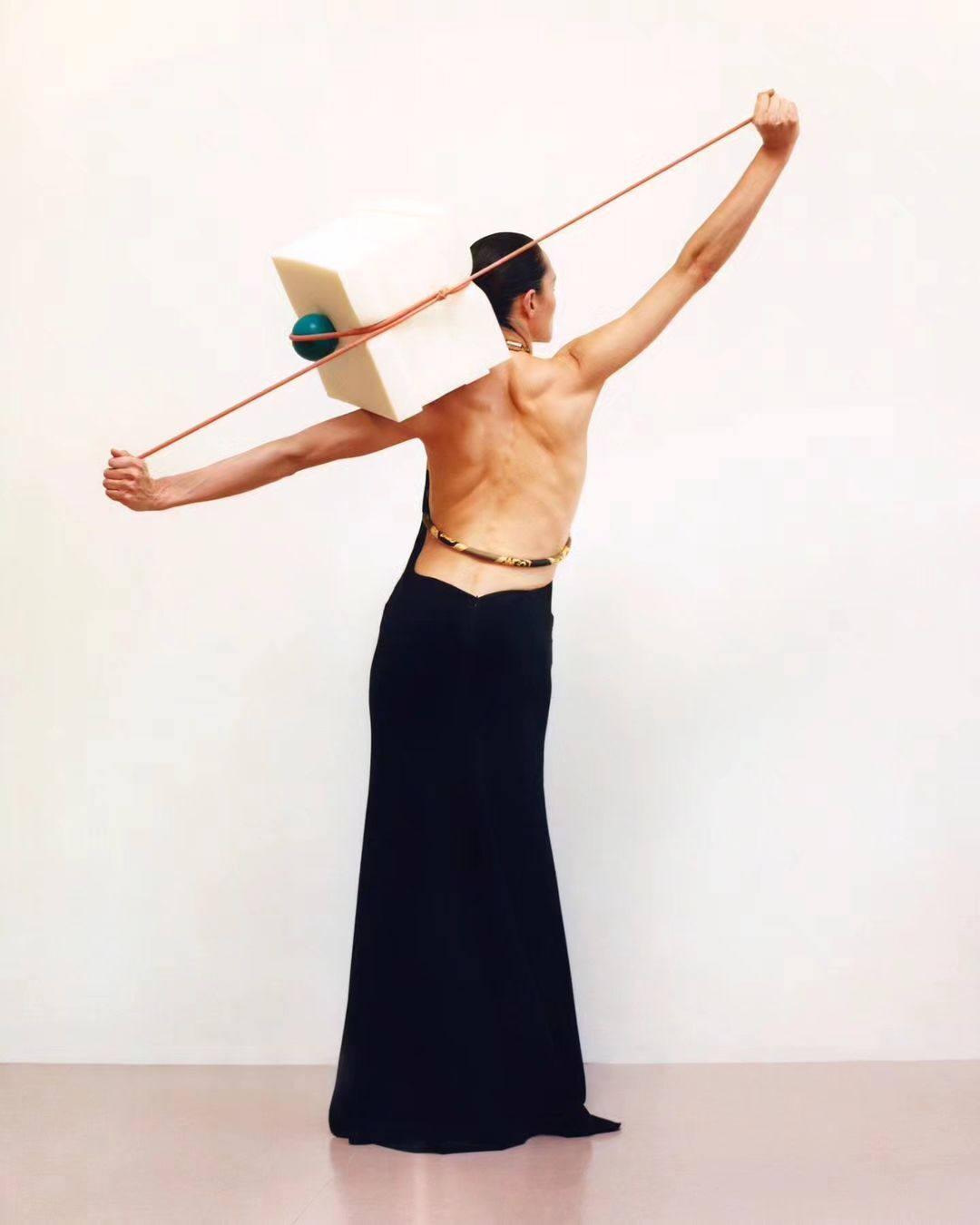 A woman in a black dress stands in a studio, holding a white cube with a turquoise sphere using ropes in a minimalist art shot.