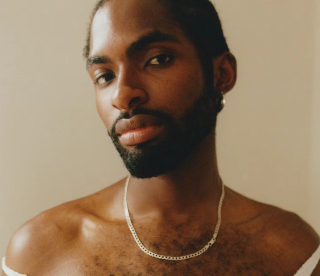 Portrait of a dark-skinned adult male with a beard, wearing a silver chain necklace, indoors against a neutral background, close-up shot.