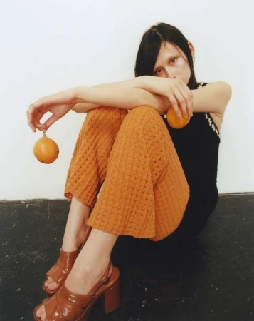 Seated woman wearing orange textured trousers and brown heeled sandals holding small round fruits against a white wall.