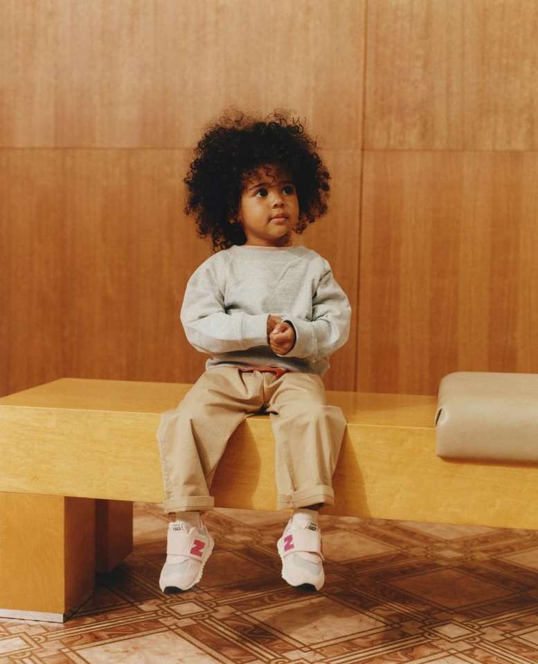 A young child with curly hair sits on a yellow bench, wearing a light blue sweater, beige pants, and white and pink sneakers in a studio setting.