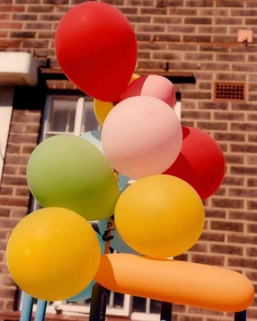 Colorful balloons against brick wall. Red, yellow, pink, and green balloons are attached to a blue metal stand outdoors.