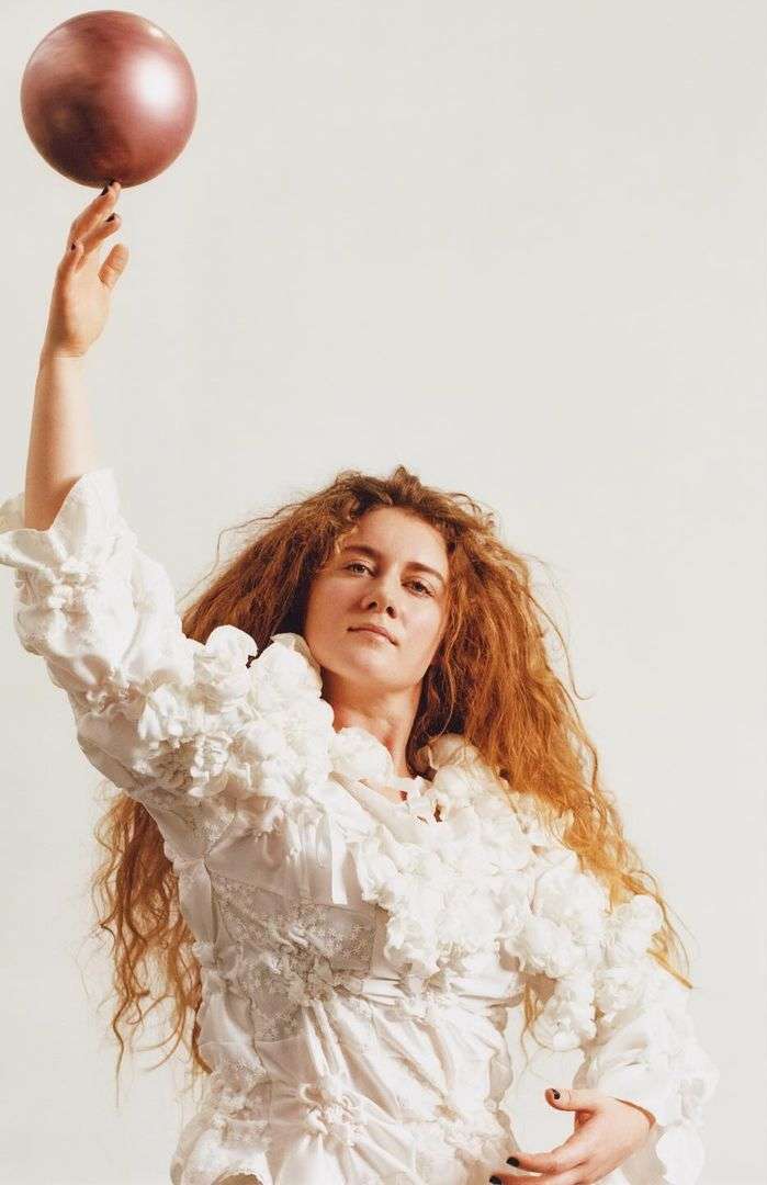 Woman with red curly hair poses with a metallic ball in a white photo studio, wearing a textured, ruffled white dress.