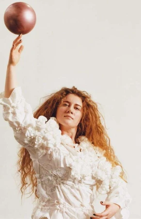 Woman with red curly hair poses with a metallic ball in a white photo studio, wearing a textured, ruffled white dress.
