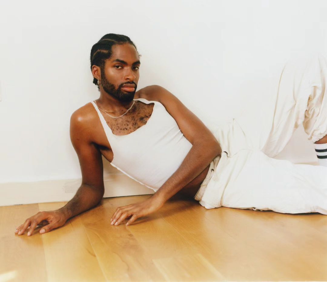Portrait of a bearded, adult Black man in a white tank top, lying on a wooden floor against a white wall in a studio setting.
