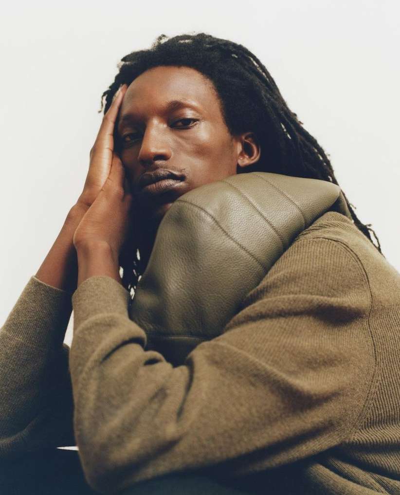 Portrait of a black man with dreadlocks wearing an olive green sweater against a plain white background.