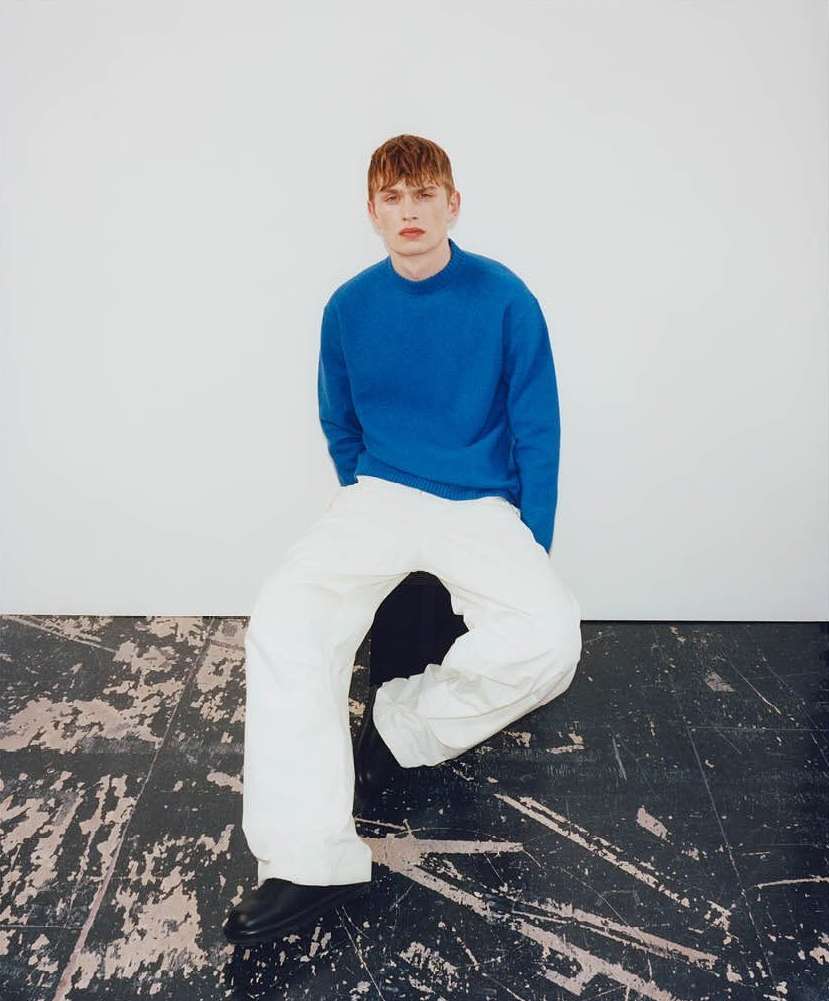 A young male model with red hair seated on a dark stool, wearing a blue sweater and white trousers against a neutral background.
