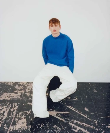 A young male model with red hair seated on a dark stool, wearing a blue sweater and white trousers against a neutral background.