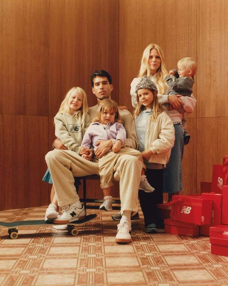 A family of six poses indoors in coordinated beige tones. Four young children and two adults wear apparel and footwear from a New Balance advertisement.