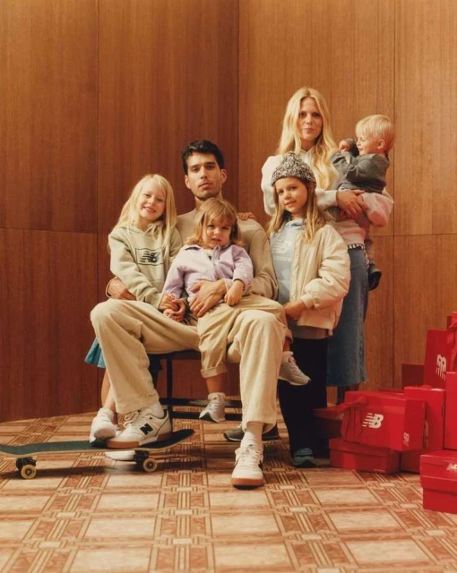 A family of six poses indoors in coordinated beige tones. Four young children and two adults wear apparel and footwear from a New Balance advertisement.