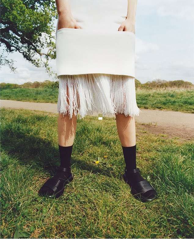 Woman in white dress with fringe detail and black sandals on grass in a field on a bright day for a fashion forward editorial.