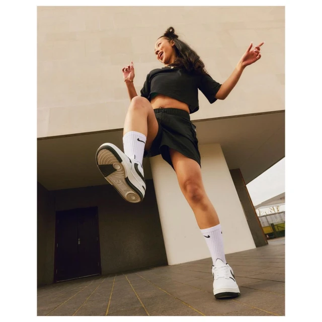 Athletic woman jumps with energy outside wearing Nike shoes, socks, and sportswear. Shot at a low angle against an architectural background.