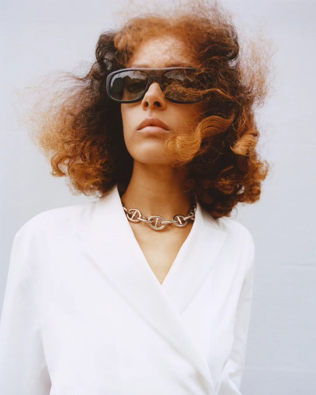 Woman with curly red hair wears sunglasses and a silver chain necklace while posing in a white wrap top against a grey background.
