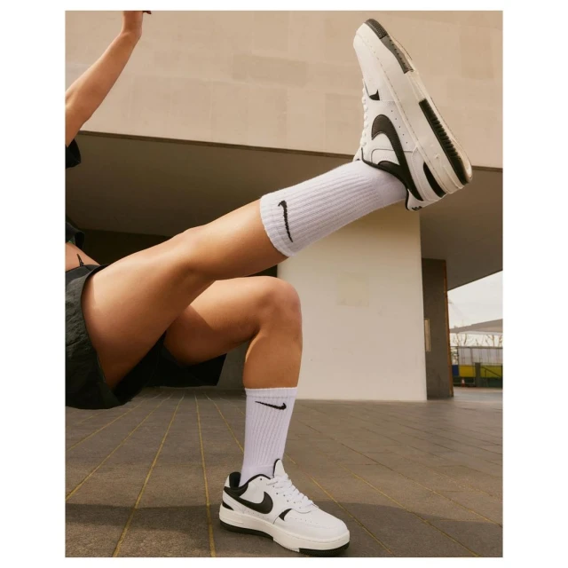 Woman with legs outstretched wearing white Nike sneakers and socks. An outdoor urban setting with a concrete floor and wall.