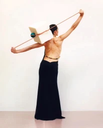 Back view of a woman in a black gown holding a taut rope balancing a white cube with a green sphere by photographer Laura Mccluskey.