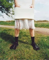 Close-up of a person wearing a white skirt with fringe, black socks, and black sandals standing on grass outdoors