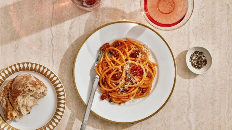 Overhead shot of bucatini pasta with tomato sauce on a white plate with a golden rim, accompanied by bread and a glass of rosé wine.