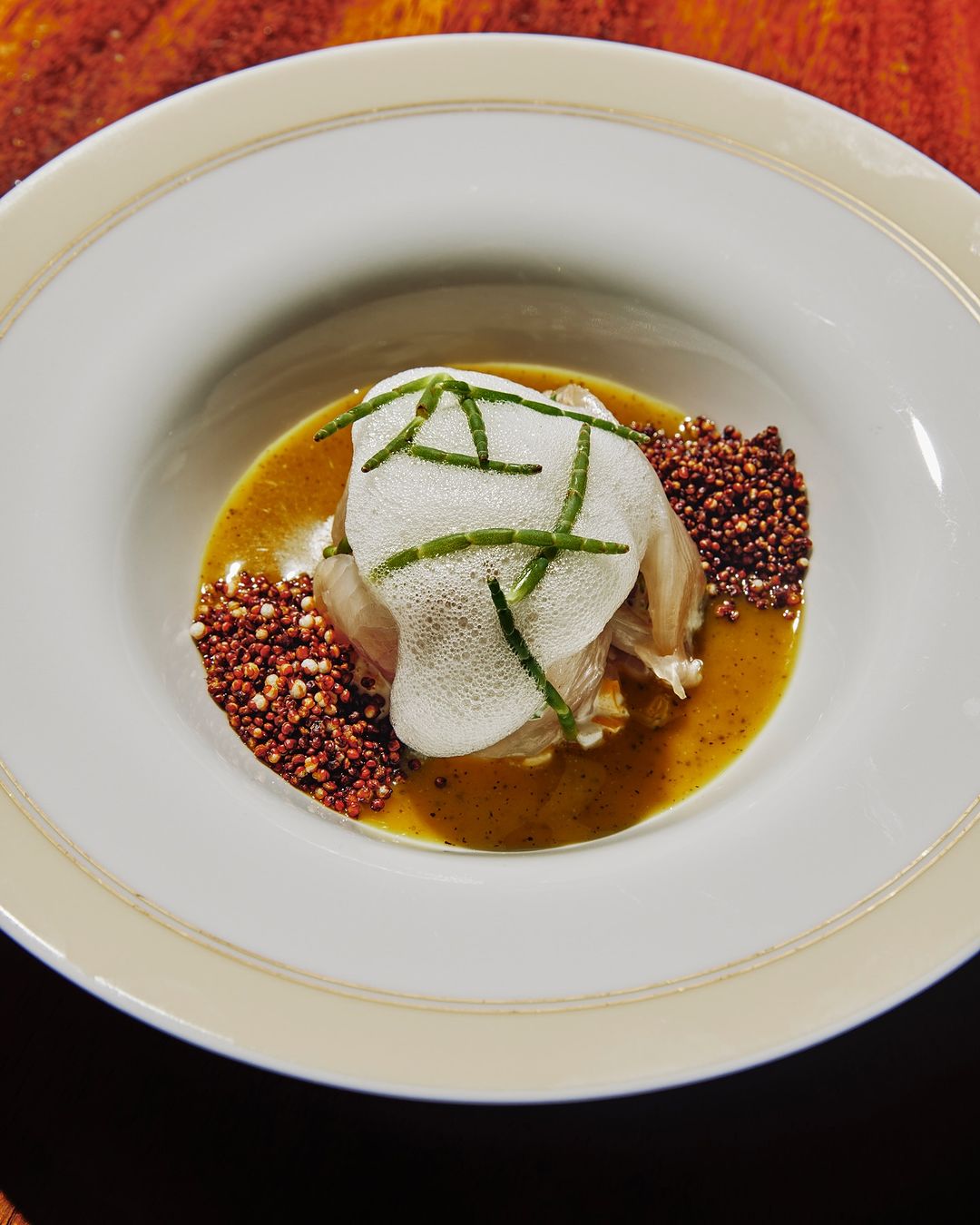 Overhead shot of a meticulously plated dish featuring white fish, foam, green herbs, brown puffed grains, and a light brown sauce in a bowl.