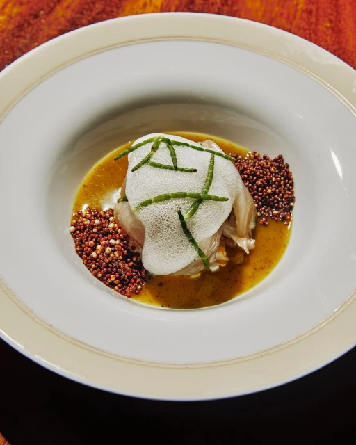 Overhead shot of a meticulously plated dish featuring white fish, foam, green herbs, brown puffed grains, and a light brown sauce in a bowl.