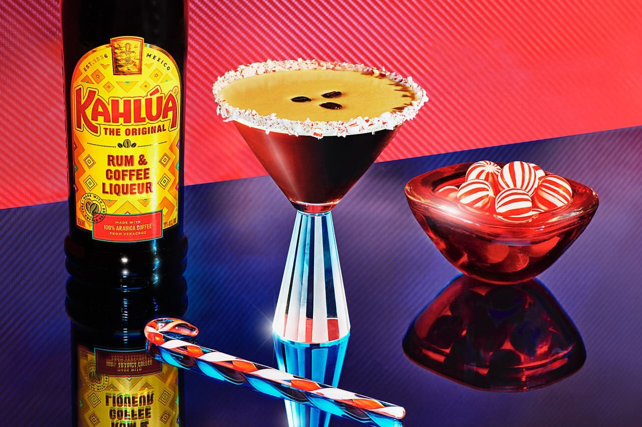 Kahlúa liqueur bottle and a martini glass with coffee liqueur, candy cane, red glass bowl with striped candies, red/blue backdrop.
