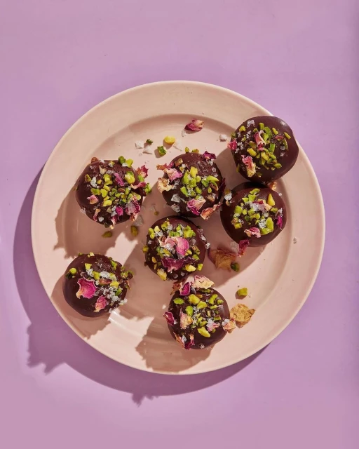 Overhead shot of chocolate dessert truffles on a pink plate, decorated with pistachios and rose petals, set against a lilac background.