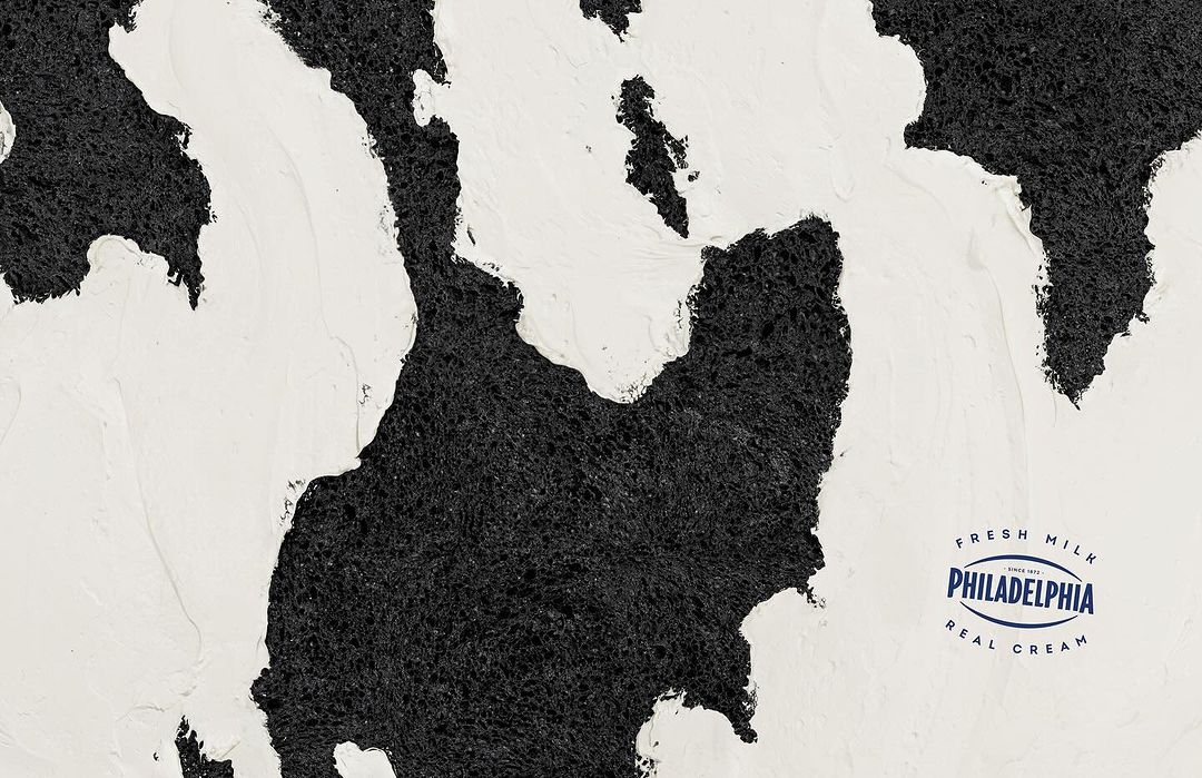 Creamy white topping swirls over a dark, textured surface, creating an abstract design with the Philadelphia Cream Cheese logo.