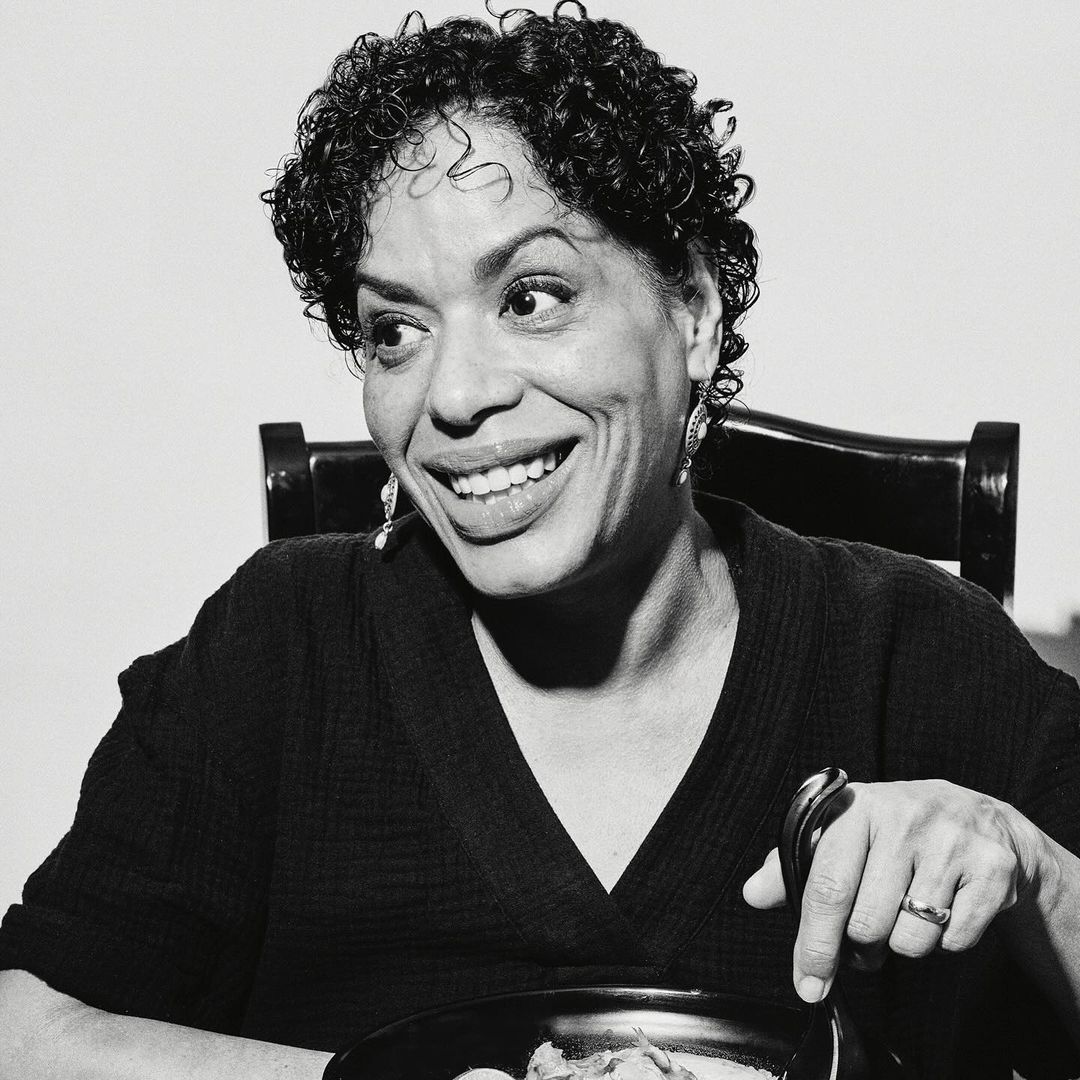 Smiling woman in black-and-white portrait sits at a table with a pot of food and a spoon, radiating joy in an indoor setting.