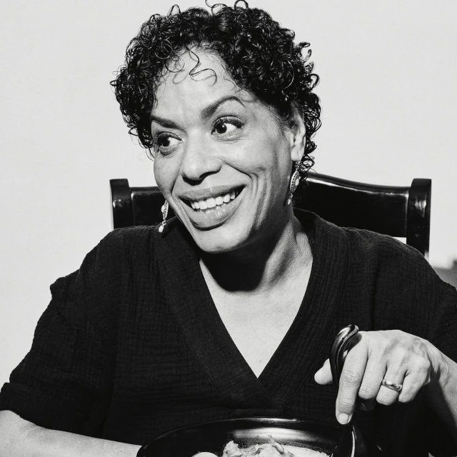 Smiling woman in black-and-white portrait sits at a table with a pot of food and a spoon, radiating joy in an indoor setting.