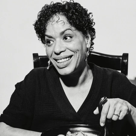 Smiling woman in black-and-white portrait sits at a table with a pot of food and a spoon, radiating joy in an indoor setting.