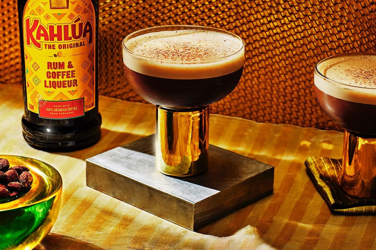 Espresso martini cocktails next to a Kahlua bottle, served on silver block. Drink photography showcases golden hues and warm lighting.