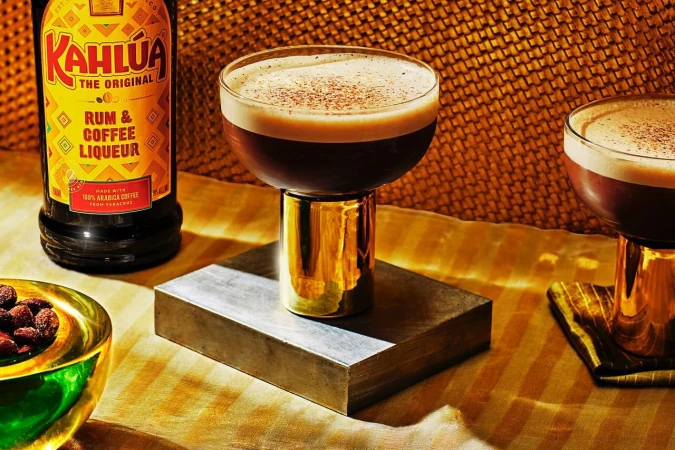 Espresso martini cocktails next to a Kahlua bottle, served on silver block. Drink photography showcases golden hues and warm lighting.