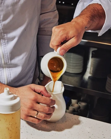 Chef's hands pouring sauce from a white pitcher into another, close-up shot in a restaurant setting with culinary preparation.