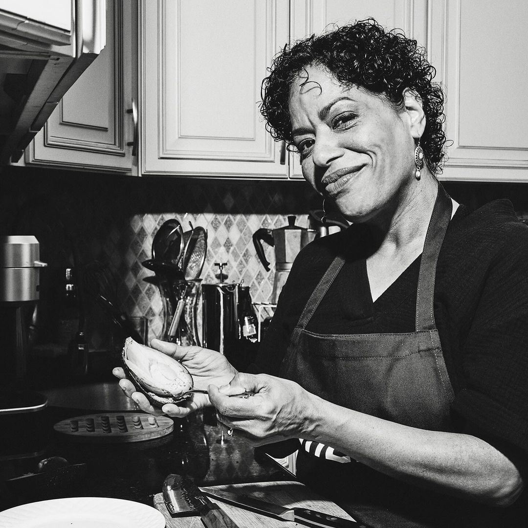 Black and white portrait of a smiling mature woman indoors wearing an apron, peeling an avocado in a kitchen with dark countertops.