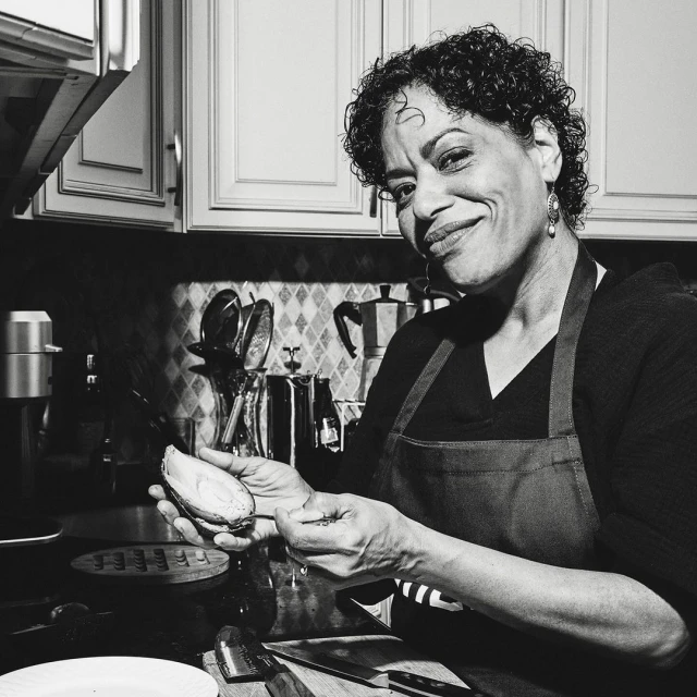 Black and white portrait of a smiling mature woman indoors wearing an apron, peeling an avocado in a kitchen with dark countertops.