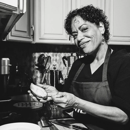 Black and white portrait of a smiling mature woman indoors wearing an apron, peeling an avocado in a kitchen with dark countertops.