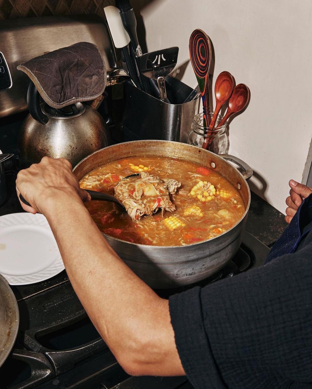 Person stirs a large pot of soup on a black stove in a kitchen with a kettle, utensils, and wooden spoons in the background.