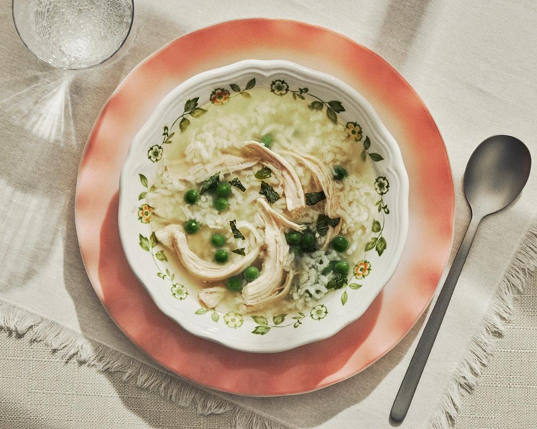 Overhead shot of chicken and rice soup with green peas in a floral-patterned bowl on a pink plate, accompanied by a spoon and a glass of water.