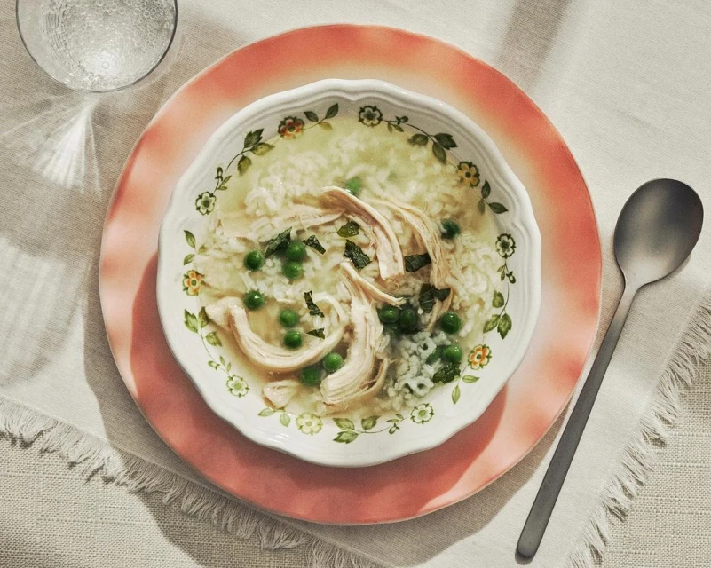 Overhead shot of chicken and rice soup with green peas in a floral-patterned bowl on a pink plate, accompanied by a spoon and a glass of water.