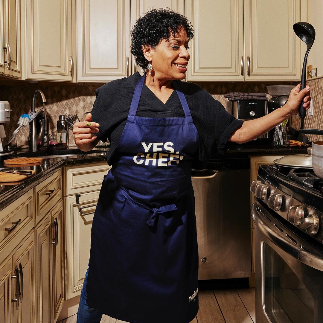 Adult woman smiles in home kitchen with a blue apron, holding a ladle, posing near a stove, with kitchen cabinets and backsplash.