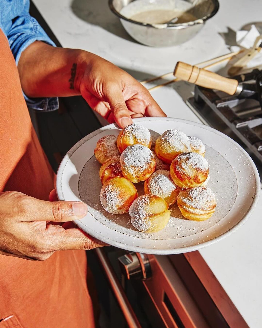 Close-up of a person holding a plate of Aebleskiver pancakes dusted with powdered sugar, presented indoors with a stove in background.