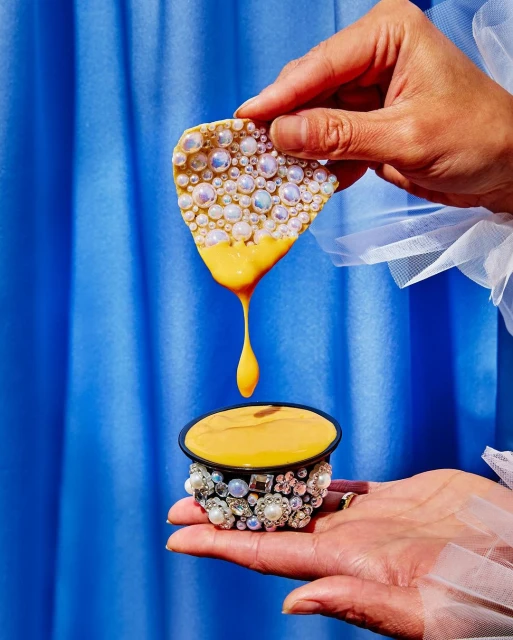 Hands holding food and liquid in a container decorated with pearls, against a blue curtain backdrop, in a studio-lit setting.