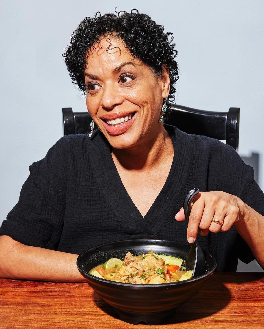 Close-up of a smiling, middle-aged woman with curly black hair preparing to eat a bowl of vegetable soup on a wooden table.