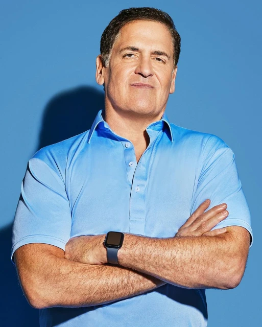 Mark Cuban poses in a blue polo shirt with arms crossed, showcasing his smartwatch against a bright blue backdrop in a studio shot.