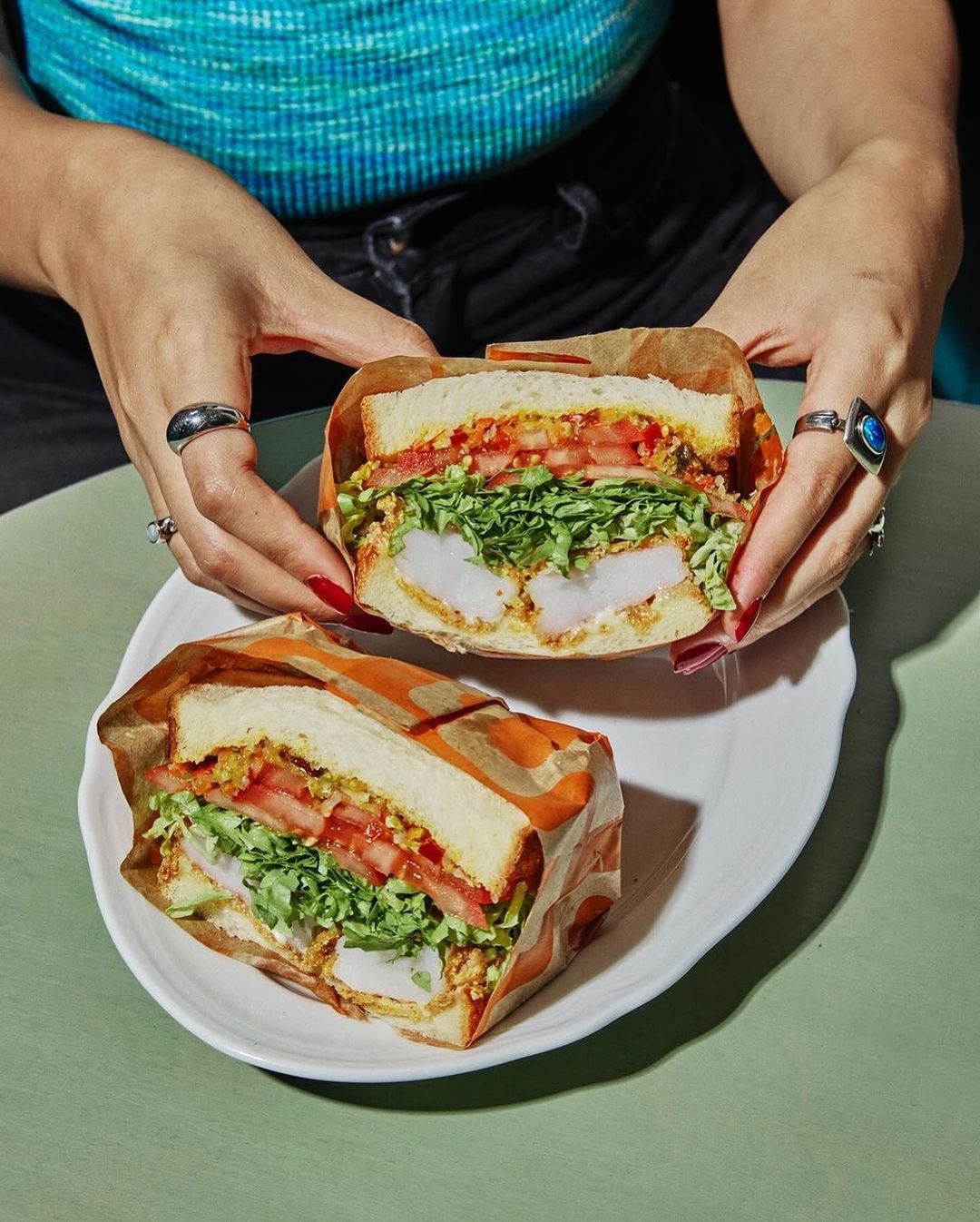 A person holding a sandwich cut in half wrapped in paper on a white plate. The sandwich contains fish, lettuce, and tomato.