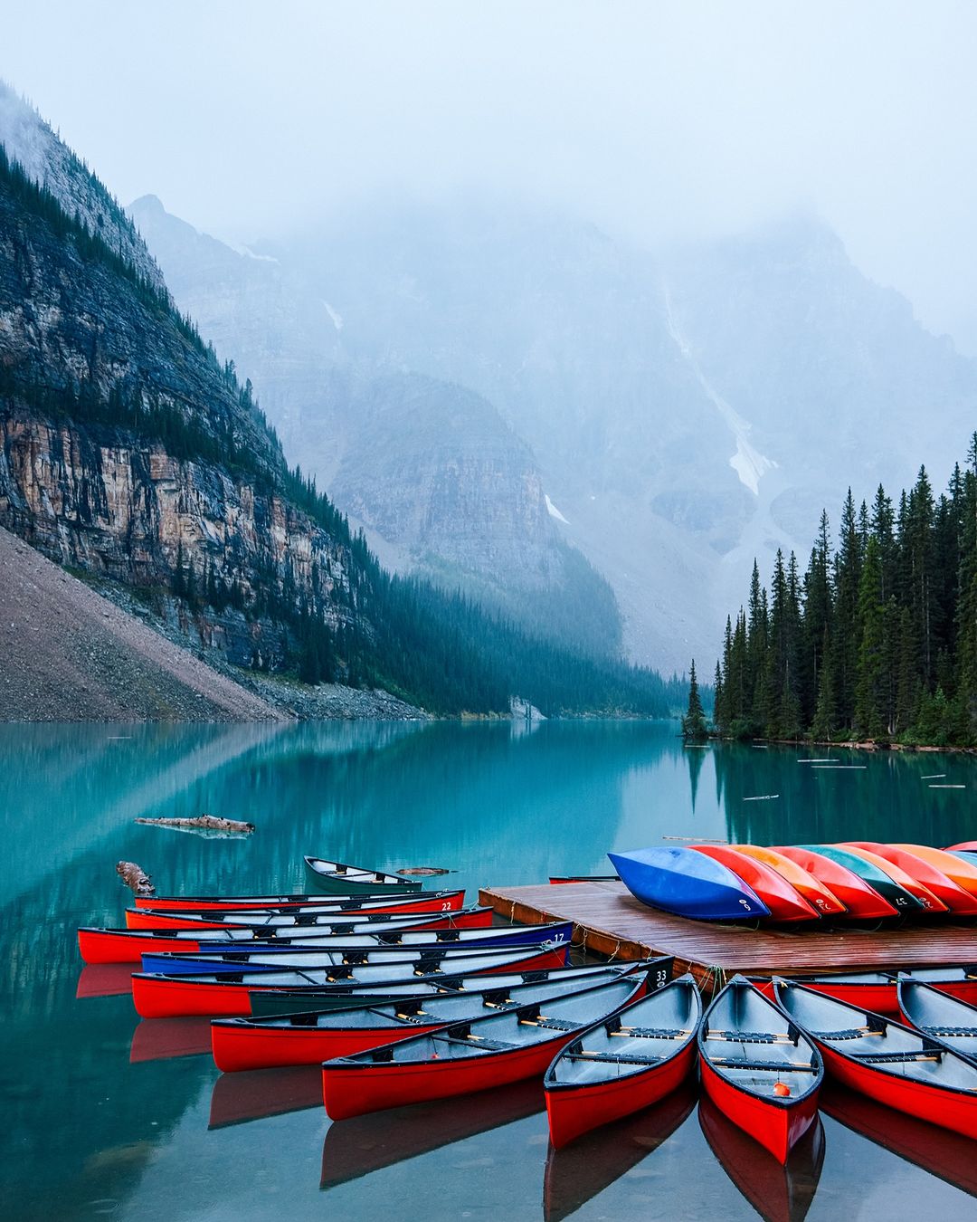 A serene view of canoes docked on a wooden pier, set against a backdrop of turquoise lake, mountain range, and dense forest.