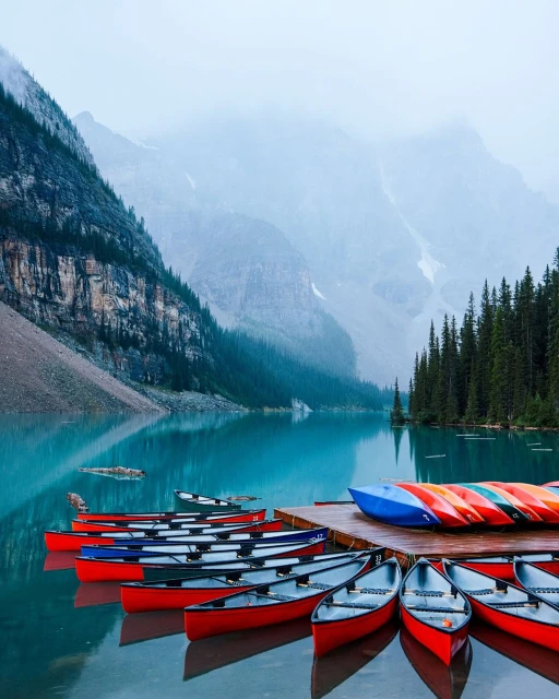A serene view of canoes docked on a wooden pier, set against a backdrop of turquoise lake, mountain range, and dense forest.