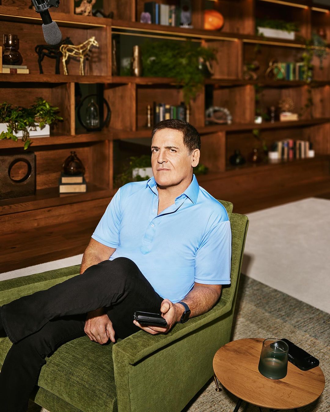 Adult male in a turquoise collared shirt sitting in a green armchair with black pants, holding a smartphone, with wooden bookshelves.