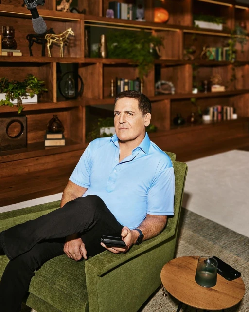 Adult male in a turquoise collared shirt sitting in a green armchair with black pants, holding a smartphone, with wooden bookshelves.