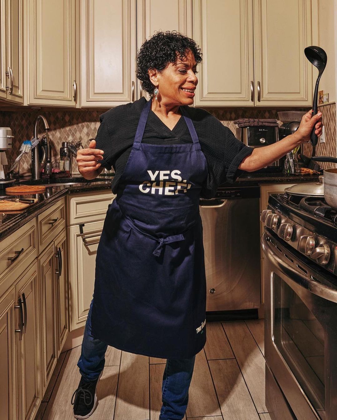 Enthusiastic African-American woman in kitchen apron strikes a joyful pose with a cooking ladle amidst light beige kitchen cabinetry.