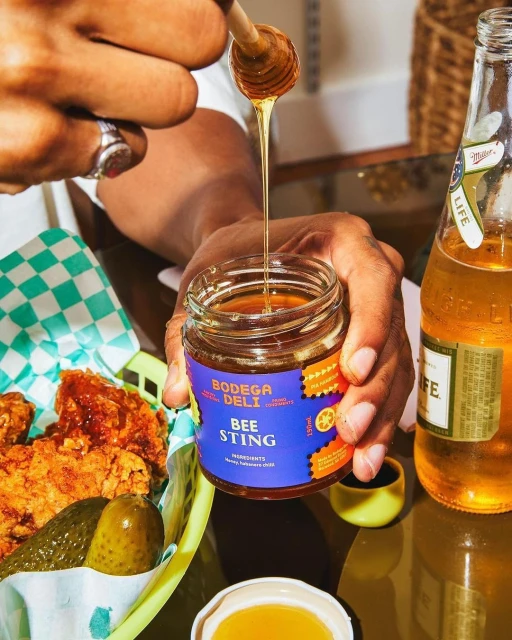 Person pours honey from a wooden dipper into a jar, featuring fried chicken, pickles, and a beer bottle in an indoor setting.