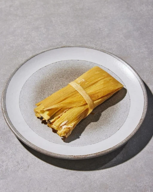 A wrapped tamale on a speckled grey plate sits on a grey surface. The tamale is bound with a thin strip of corn husk.